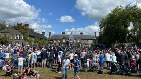 A large group of spectators standing on grass in front of a large house