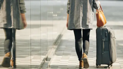 Getty Images A woman walks a suitcase down the street with her back to the camera.