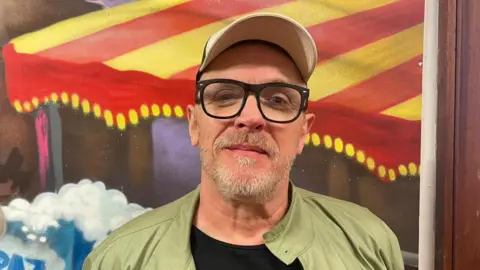 A head-and-shoulders shot of a man with a greying closely shaved beard in cap and black-framed glasses. he is wearing a khaki jacket and black t-shirt and standing in front of a painted backdrop. Painting in the background depicts a red and yellow striped canopy.