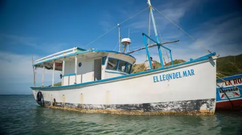 A small white boat with blue trim is seen at anchor. The boat is called "El Leon del Mar".