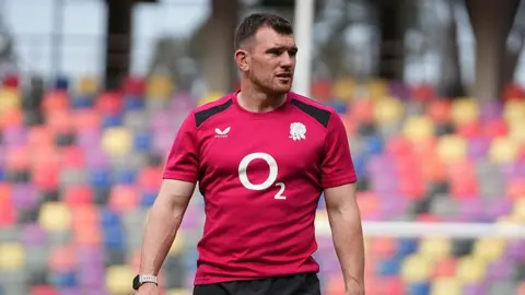 Ben Spencer is wearing a red England training shirt and standing on the pitch during a pre-match warm-up ahead of the game with Argentina in July.