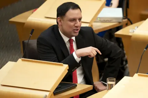 PA Media Anas Sarwar wears a dark suit and a red tie. He is mid-conversation while sitting in the debating chamber in Holyrood.