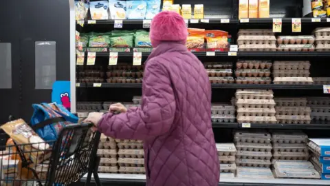 Getty Images Uma mulher em um casaco inchado roxo empurra um carrinho de compras passando por ovos no supermercado