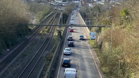 The A48 road pictured from a bridge above, showing cars driving along a road which runs adjacent to a train track.