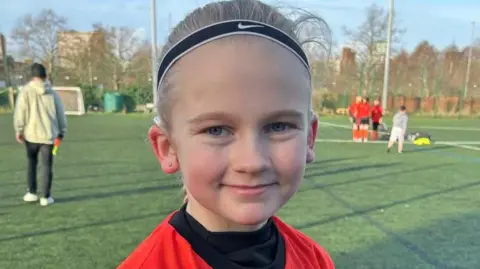 Catherine Souster Milly smiles for the camera while on the pitch on a sunny day. Other players can be seen behind her.