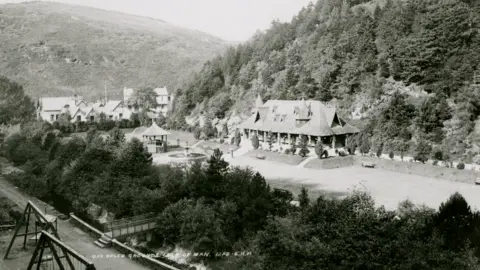 Manx National Heritage An early 20th Century black and white photograph showing a line of trees at the bottom, with a Swiss looking long cottage with pointy dormers raised over a long lawn, with a fountain in front and a gazebo. Behind the cottage is a hillside covered in trees, and there are further buildings in the distance on the left, in front of another hillside.