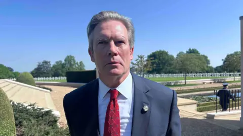 KATE BRADBROOK/BBC A man in a smart navy suit with a red tie looking at the camera. Behind him in the distance there are rows of white crosses marking the graves of servicemen. 
