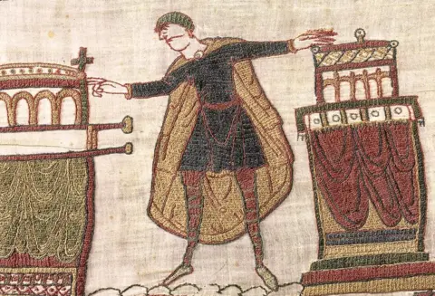 Bayeux Museum Harold depicted in the tapestry, holding arms out and touching two boxes, close cropped on the scene with fabric texture detail.