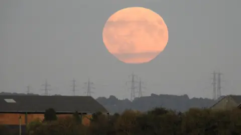 BBC Weather Watchers/Marvo A coppery big moon hovers above several pylons. Two buildings can be seen on the left and right of the picture, separated by trees and bushes.