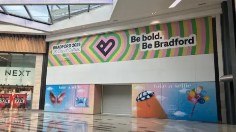 An empty shop in a large empty shopping centre. It is covered in hoarding advertising Bradford 2025 UK City of Culture
