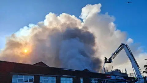 A factory building is visible with plumes of smoke billowing above it into the sky. there is a white aerial platform with a firefighter on it, and they are spraying the fire with a hose