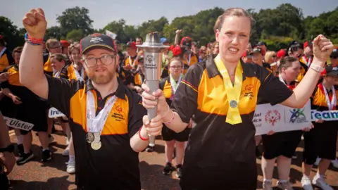 PA Media Jenna Thevanau (right) holding a Special Olympics flame with another male athlete. Both are holding their other hand in the air.