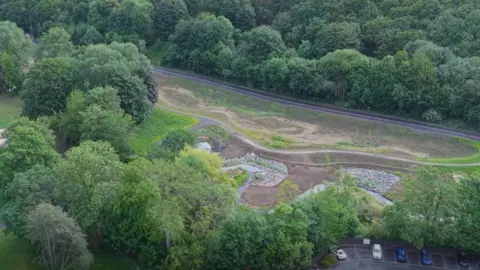 Aerial view of a landscaped wetland or flood management area surrounded by dense green woodland. Curving earth paths and shallow water channels weave through the site, with sections of exposed soil and rock-lined embankments visible. A road runs along the upper edge of the image, and a small car park with several vehicles is partially visible at the lower right.