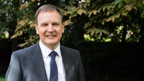 A man with short brown hair smiling into the camera from the left. He is wearing a dark grey blazer, white shirt and navy and white spotted tie. Behind him are the leaves on a tree which is blurred.