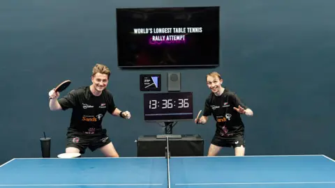 Tom Lewis Image of Dan Ives and Lloyd Gregory. They are by a table tennis table and a clock reading 13 hours 36 mins 36 secs. 