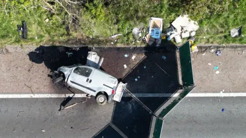 Lancashire Police An aerial drone shot of the van, lying on its side on the hard shoulder of the motorway. Its bonnet is badly damaged and the passenger side front wheel has been torn off. There is other debris littering the grass verge, and a screen has been put up around the scene.