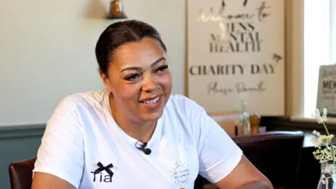 Tia Davies wearing a white t-shirt with her name on it. She is smiling and looking to the side of the screen.