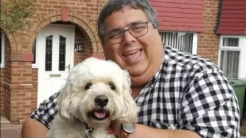 Councillor Richard Udall stands in a garden outside a red brick property. He wears glasses and a short-sleeved checked shirt. He is holding his white dog which looks like a poodle-cross.