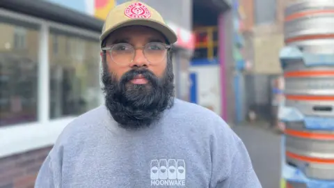 Vinny Rosario looking straight at the camera. He is wearing a grey jumper with a white Moonwake logo on it. He has a large black beard and glasses and is wearing a tan-coloured hat with a red logo on it. He is standing in front of a blurred background with a stack of kegs.
