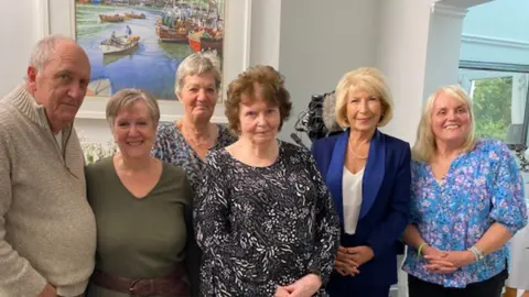 Jane Jackson Six grandparents, including Jane, standing in a home together in a group, all looking to camera with a picture of a river and boats on the wall behind. Jennie Bond, the former BBC journalist and television presenter, is amongst them, wearing a blue trouser suit and string of pearls