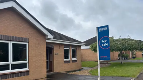 Simon Thake A smart brick bungalow with a neat grass lawn and a For Sale sign in the driveway
