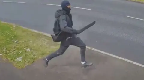 man wearing all back runs across grass verge holding a machete