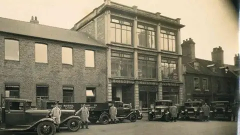 Gerald Dover Thomas Green A historical black and white image of 33-39 St James Street in King's Lynn. In front of the building there are six old-fashioned cars and their drivers standing next to them.