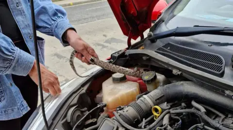 Dave Bigsby A patterned snake is stretched across the open engine bay of a car. Two hands carefully hold the snake near the front, above visible engine parts and fluid containers.