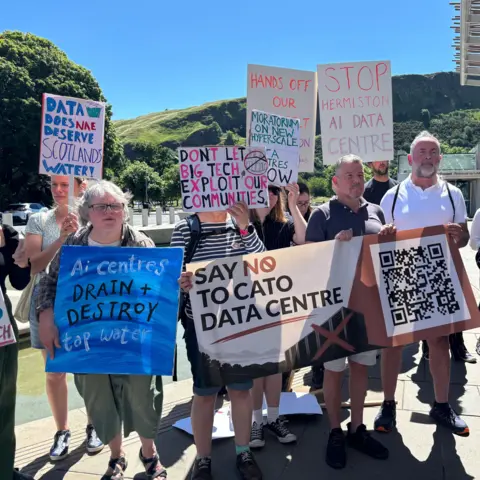 A group of people stand outdoors in bright sunshine holding protest signs opposing a data centre development. The group is gathered near a modern building with a metal canopy, with green hills and a clear blue sky in the background.