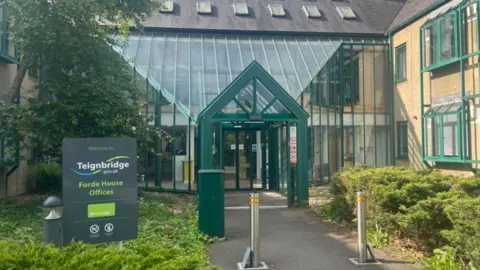 The front entrance of Forde House with a glass atrium. There are two metal bollards on the pathway to the entrance and a sign to the left.
