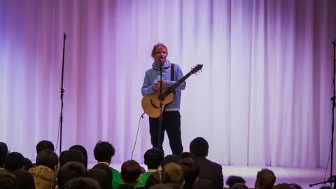 Ed Sheering on stage in front of school pupils. he is holding a guitar and is wearing a lights blue hoodie and dark trousers