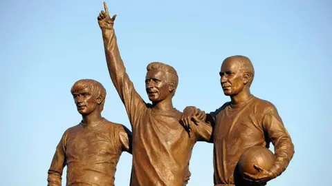 Getty Images A bronze statue of three men in football strips. The one in the middle raises his hand to point at the sky and the man on the far right holds a football.