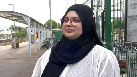 Faatimeh Bham wearing a head scarf and glasses and a white blouse in front of a railway platform.