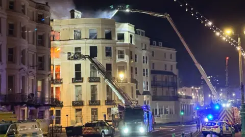 Getty Images The Royal Albion Hotel at night in Brighton in 2023. Flames and smoke are coming from some of the windows. Multiple height fire appliances are spraying water on the flames.