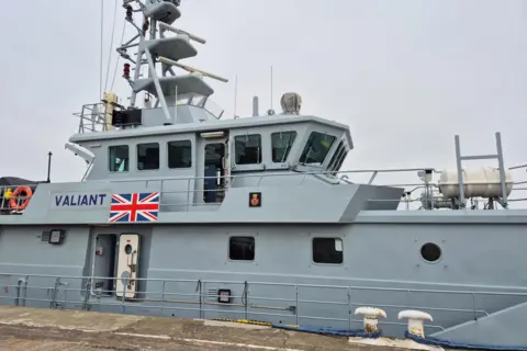 BBC / Natalie Glanvill A large grey boat at dockside with a Union Jack on the side next to its name Valiant. There is equipment on board including a life ring. There are windows and two doors, one which is open. The boat is moored to the dock with blue ropes.