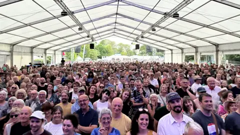 Hannah Olsson/BBC A large crowd of people all facing the stage at the folk festival. The camera has been positioned on the stage so all of their faces can been seen. They have gathered under a large white tent and many people are smiling. 