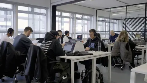 Hospital records employees sitting at gray desks in a warehouse-style office with their laptops. About seven members of staff are visible, young people wearing hoodies and T-shirts mostly in monochrome tones; Some have thick headphones. Last but not least is founder Chris Goss, an older man with gray hair, a Crittall-style glass screen and a black-and-white abstract mural.