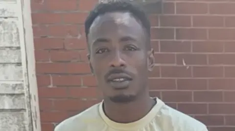 A man in a light yellow t-shirt stands looking forward with a brick wall behind him. 