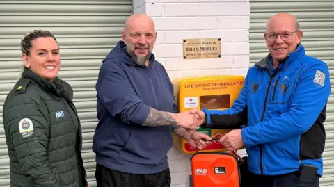 Three people standing in front of the yellow defibrillator box installed on the wall. There is a woman wearing a green ambulance uniform on the left. Paul Downton, in the middle, is shaking hands with William Morley on the right. They are also both holding the orange defibrillator bag. All three are smiling at the camera. A plaque above the wall-mounted box reads: "In loving memory of Billy Morley".