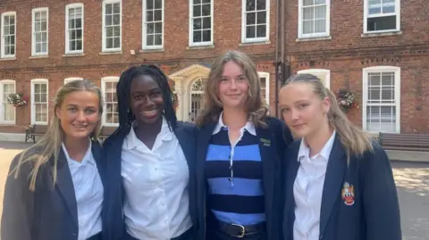 Four girls wearing blue school blazers stand in a row in front of a large brick building with large white frame windows.
