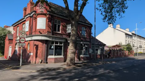 A red-brick pub on the corner of a road, with a tree in front of it.