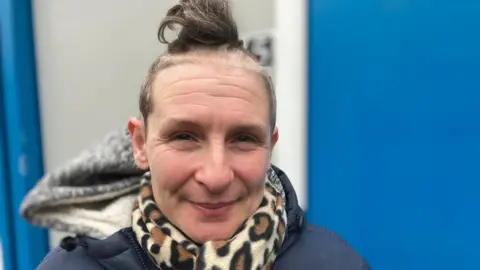BBC Emma is standing outside her pod. She has dark hair tied up and is wearing a blue rain jacket, a grey hoodie and a leopard-print scarf.