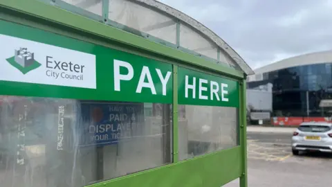 A plastic shelter which houses pay and display machines has an Exeter City Council logo on the left and pay here written in capitals