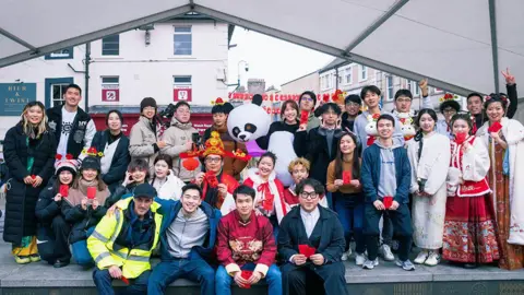 Popber and Edward Au Dozens of organisers, performers and volunteers of the Chinese New Year festival gathered on the stage. Some are wearing traditional Chinese clothing and costumes. They are all holding traditional Chinese red envelopes.