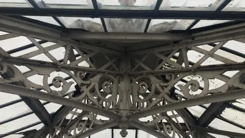 Andrew Turner/BBC A close up picture of the ornate iron work within the top of the lantern of the Great Yarmouth Winter Gardens, showing the grey sky through dirty glass