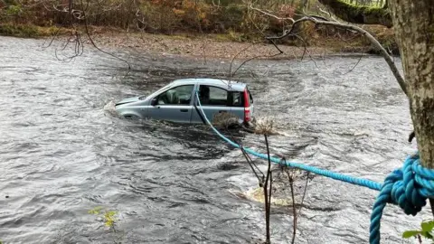 BBC A car partially submerged in a fast-flowing river, secured by a blue rope tied around a tree in the foreground, with bare trees and grassy banks in the background.