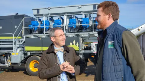 DAERA Andrew Muir with Jack Blakiston Houston from BH Estates. Andrew Muir is wearing a brown coat, light blue shirt, yellow bow tie and glasses, and has his hands together. Jack Blakiston Houston has ginger hair and beard, wearing a blue puffer vest and green jumper with his arms behind his back. Farm equipment is seen in the background.