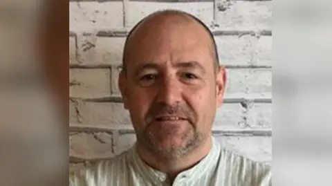 Neath Port Talbot Council A head shot of Lewis from the council website. He has short, cropped hair and a stubble on his face. He is wearing a beige, round-neck button-down shirt with white vertical stripes. The background is a grey, brick wall patterned wallpaper. 