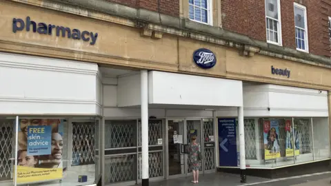 David Freezer/BBC A Boots pharmacy and retail shop is pictured with shutters across its doors as a customer reads a notice explaining its closure in a window