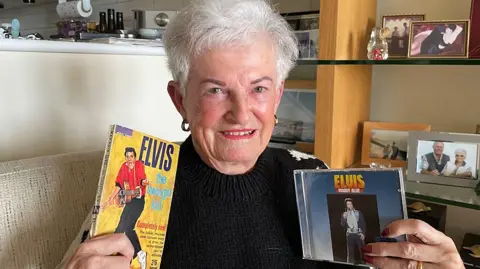 Jan Owen, an older woman, who is sitting indoors, smiling warmly while holding Elvis Presley memorabilia. In her right hand, she holds a book featuring Elvis on the cover with the text "Elvis: The Swingin’ Kid." In her left hand, she holds a CD case titled "Elvis: Moody Blue." The background includes shelves with family photographs and decorations